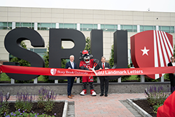 Two men in suits stand next to mascot and cut red ribbon.