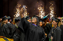 A graduate takes a selfie in a crowd of fellow graduates as sparklers light up the background.